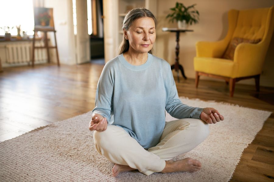 Calm barefoot middle aged female sitting on carpet in half lotus posture, making mudra gesture and closing eyes, having peaceful facial expression, doing mindful meditation, concentrating on breathing
