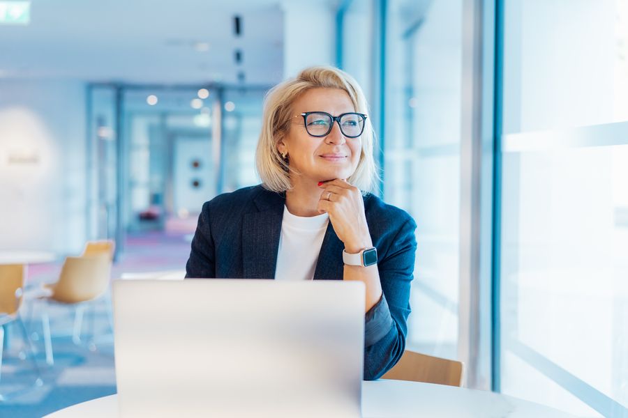 50's confident mature businesswoman dreamy looking at window, middle-aged experienced senior female professional working on laptop in open space office. Female entrepreneur working remotely